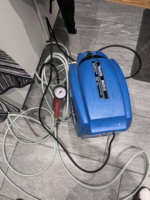 Luftkompressor och tryckmätare på ett grått klinkergolv, används för att fylla på däckluft vid pyspunka.