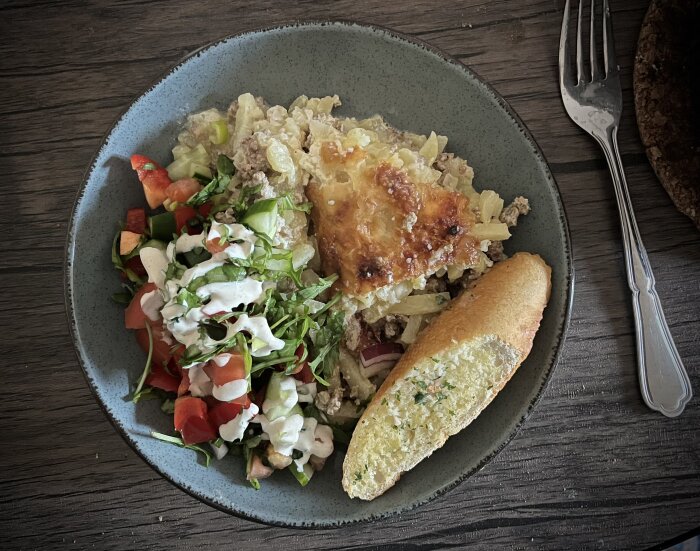 Köttfärsgratäng på tallrik med osttäcke, sallad med dressing och vitlöksbröd, serverat på ett träbord med en gaffel bredvid.
