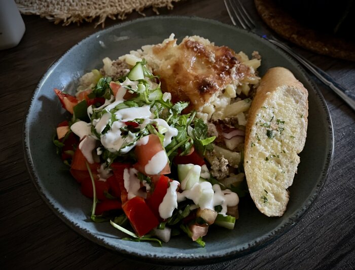 Tallrik med köttfärsgratäng, sallad med dressing och vitlöksbröd på ett mörkt underlag.