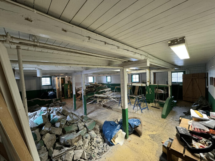 Inomhusvy av gammal ladugård med raserat material och synliga träbjälkar. Pelare stöttar taket, olika verktyg och stökigt arbetsutrymme syns.