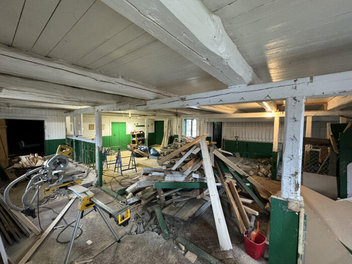 Snickeri under renovering i en äldre ladugård, med synliga bjälkar, pelare och byggmaterial. Verktyg och bråte på golvet.