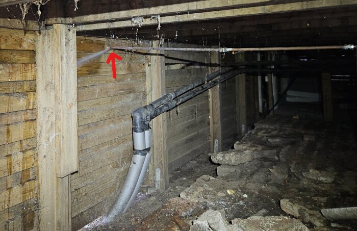 Bild av en läckande kopparrör i krypgrund under ett trähus med en röd pil som markerar brottet där röret har frusit sönder.