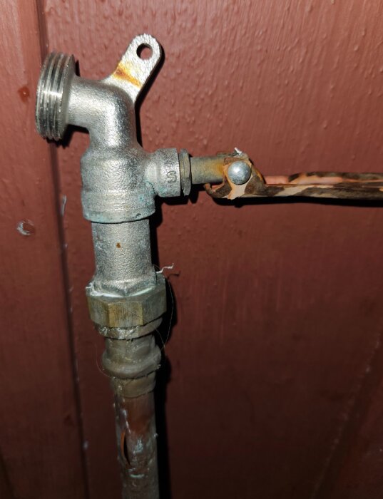 Utomhuskran med kopparrör och klämringskoppling, tydliga rostskador på röret. Röret har en läcka vid ett böjt parti framför en brun trävägg.