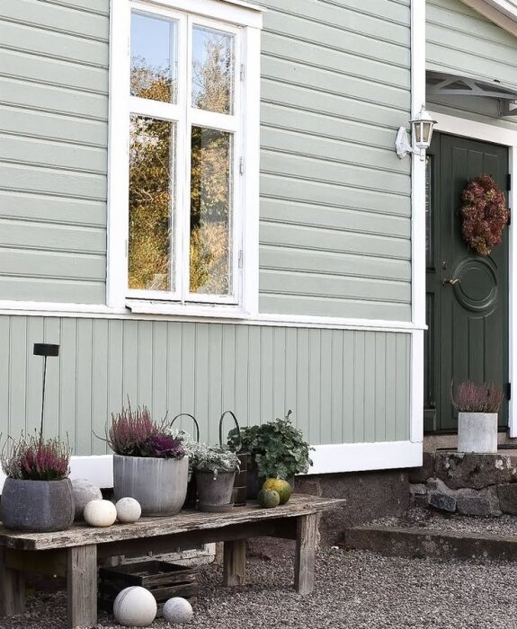 Sekelskifteshus med ljusgrön fasad och bröstning under fönster. Krukor med växter och dekorationer på bänk framför huset.