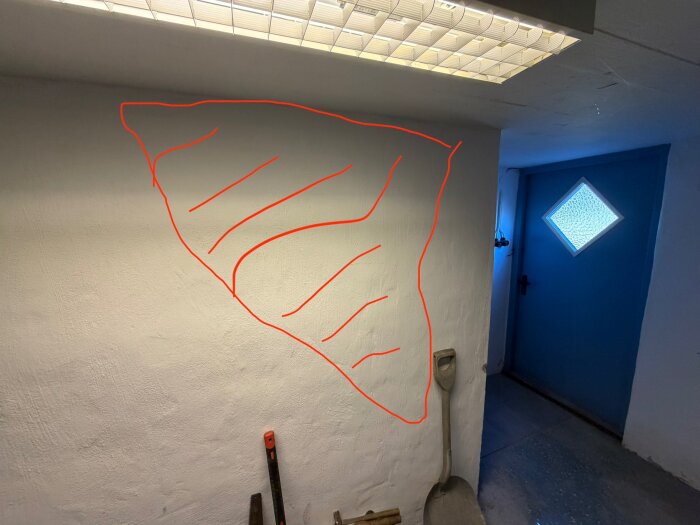 Vägg i källare med markerad sektion för planerad öppning. Röd linje indikerar var väggen kan öppnas. Blå dörr och spade syns i bild.