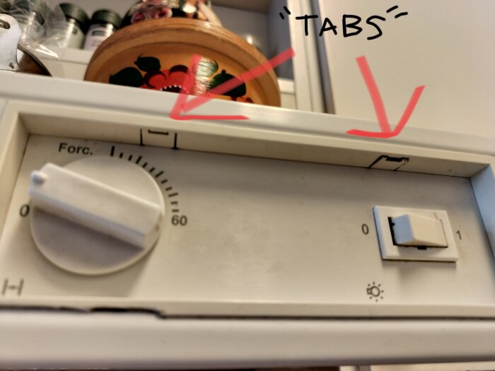 Spiskåpa med timer och inställningsvred, röda pilar pekar på två metallflikar ovanför kontrollpanelen.