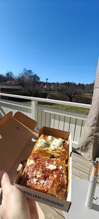 En hand håller i en pizzakartong med två bitar pizza utomhus på en altan med utsikt över en trädgård och klart blå himmel.