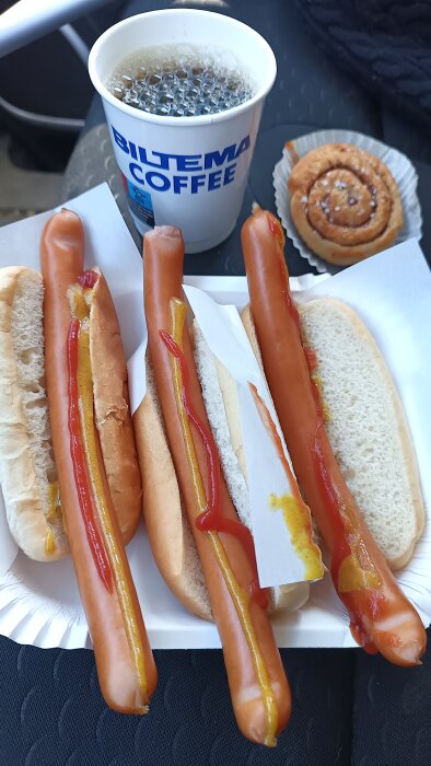 Tre korv med bröd med senap och ketchup, en kopp Biltema kaffe och en kanelbulle på ett bord.