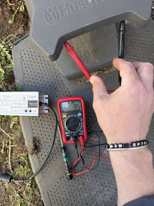 Hand mäter spänning med en multimeter bredvid en elektrisk enhet och tråd på en gummimatta utomhus.
