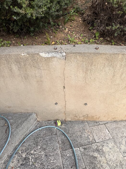 Sprucken mur vid foten av en grönskande slänt, med några skruvar i väggen och en slang och en tennisboll på stenplattorna framför.