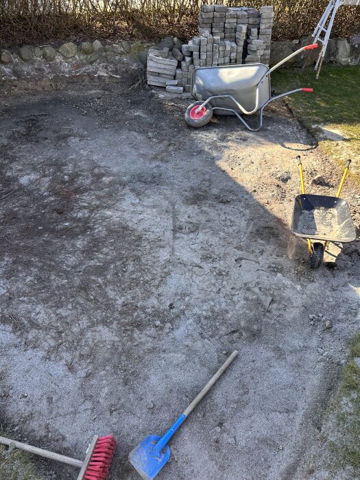 Trädgårdsområde med grus och verktyg; skottkärra, stenplattor och redskap förbereder inför gräsläggning.