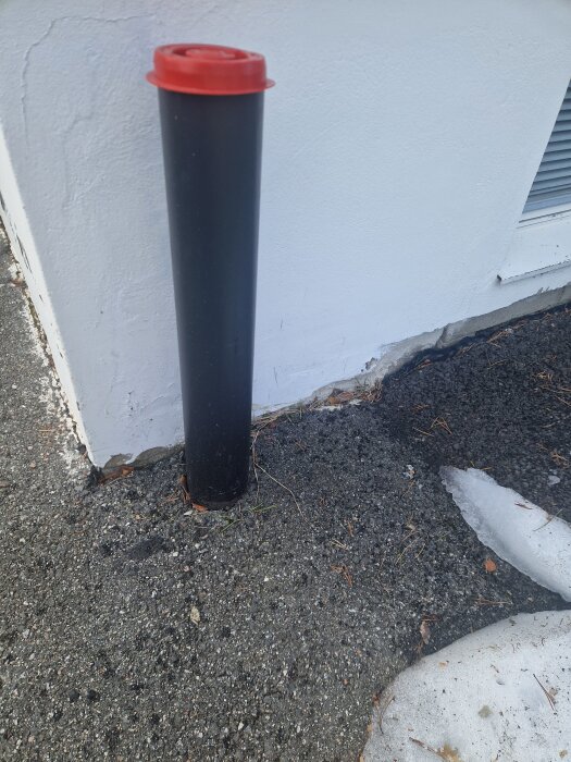 Svart dräneringsrör med rött lock vid vit källarvägg; flagnad färg syns i hörnet där trottoaren möter huset, omgiven av smältande snö.