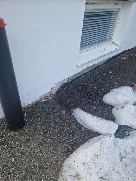 Vitt hus med källarfönster, avflagnad färg vid grundens hörn. Smältande snö vid källaren på asfalt med små rännilar av vatten vid källarväggen.