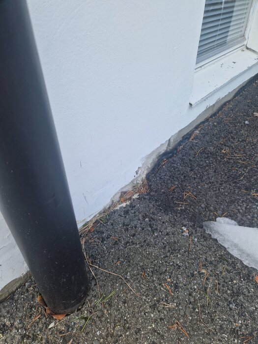 Fasad hörn med flagnande färg vid marknivå, rännsten och snö på asfalt, potentiell fuktskada nära fönster.