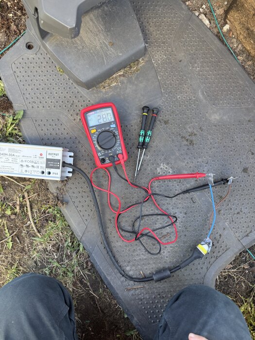 Multimeter visar 28V på lågspänningskabel vid felsökning av laddningsproblem, med verktyg och kablar på gummimatta.