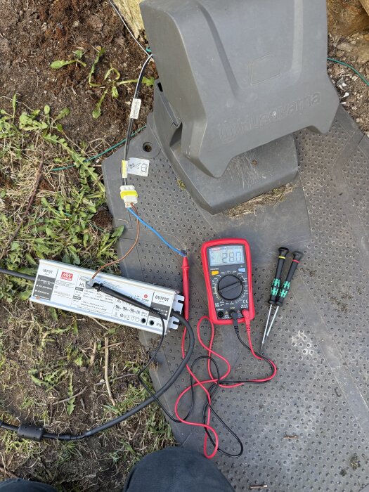 Multimeter visar 28v bredvid robotgräsklipparens laddstation, omgiven av lågspänningskabel, verktyg och växtlighet under felsökning.