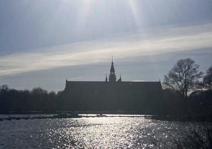 Siluett av en byggnad med torn vid en sjö, där solen glittrar i vattnet och träden syns i bakgrunden under en molnig himmel.