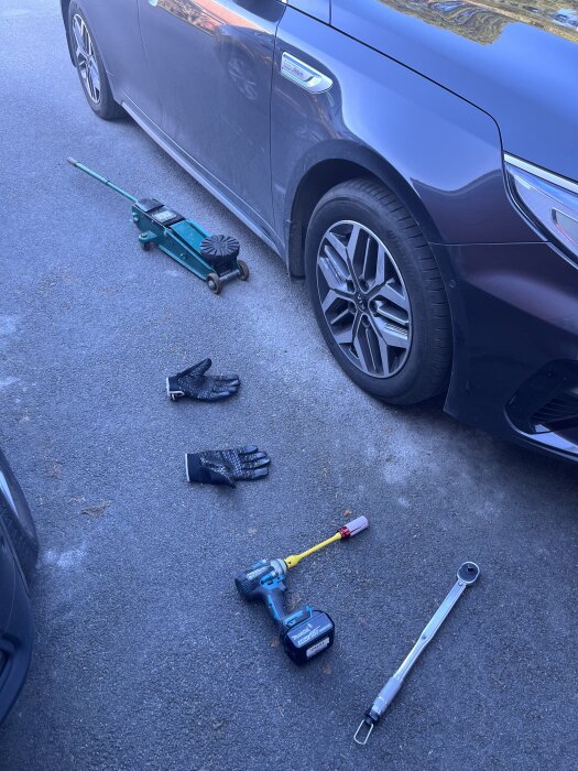 Bil, domkraft, handskar och verktyg på marken bredvid ett hjul efter däckbyte.