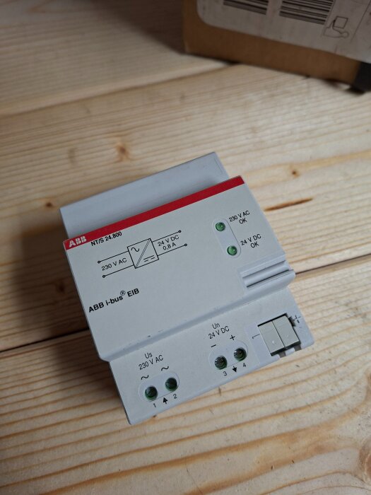 ABB i-bus EIB nätaggregat på ett trägolv, märkt med 230V AC och 24V DC specifikationer, med gröna indikatorlampor och anslutningsportar.