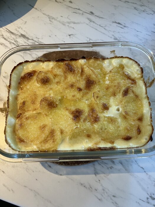 Potatisgratäng med gyllenbrun yta i en rektangulär glasform på ett marmorerat bord.