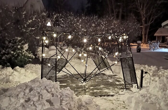 Trasig trädgårdspaviljong med hängande ljusslingor på snötäckt mark under natten.