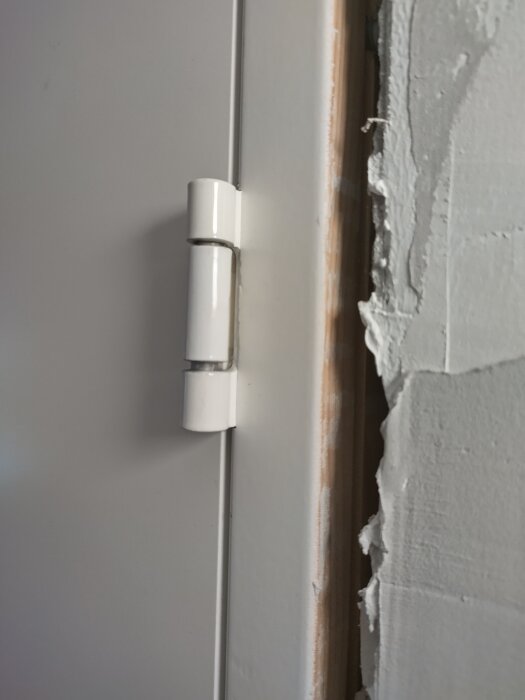 Närbild på ett gångjärn på en vit dörr, där dörrkarmen visar tecken på spackel och gips arbete.