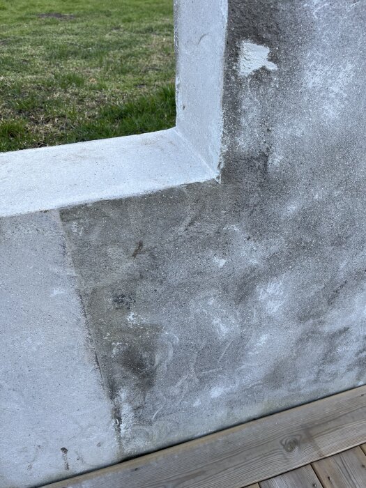 Betongvägg med rektangulär öppning och tydliga strukturer och färgskiftningar, omgiven av trätrall och grön gräsmatta i bakgrunden.