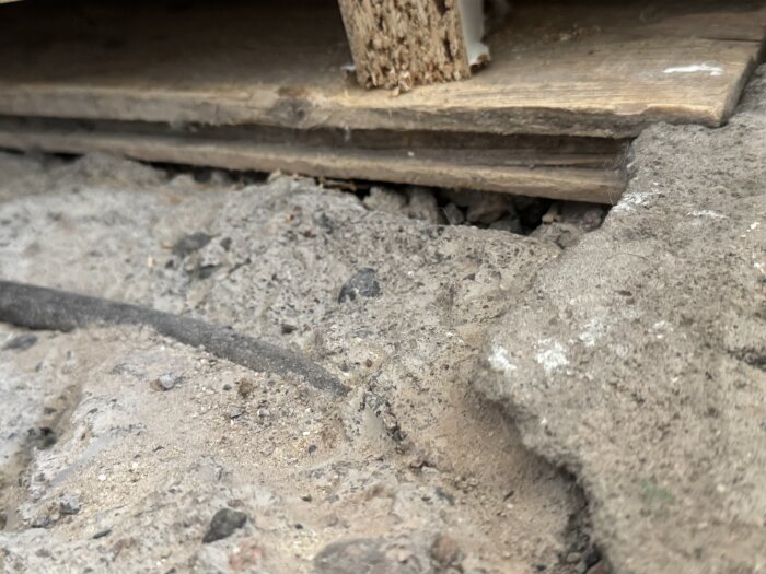 Hål i golv med synliga gamla fyllningsmaterial som sand och lera, samt en svart kabel under trägolvet i en äldre byggnad från 1915.