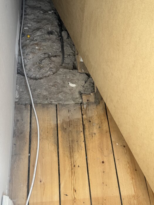 Gammalt trägolv med hål och synlig fyllning bestående av sand/lera samt en svart kabel under brädorna.