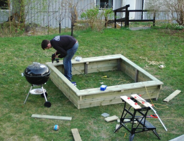 Man bygger en sandlåda av trä på en gräsmatta, omgiven av verktyg och material, med en grill intill.
