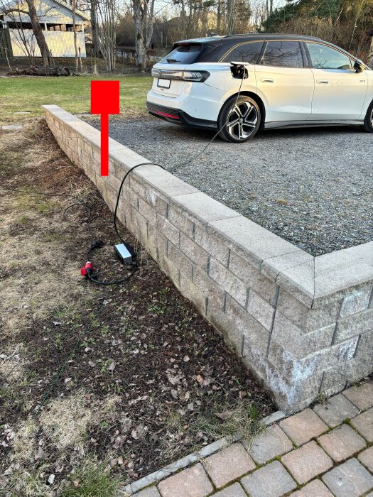 Vit elbil ansluten till en laddbox med kabel vid stenmur; muren har en toppsten som sticker ut och en röd pil markerar plats för laddboxmontering.