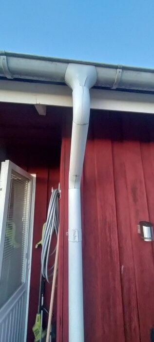 Stuprör på röd vägg vid altan, med tillhörande takränna. En upphängd slang och ett fönster syns också.