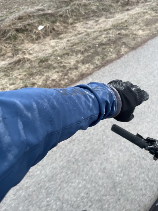 Hand i svart handske sträcker ut armen i blå jacka, cykelstyre syns, grusväg och gräs i bakgrunden.