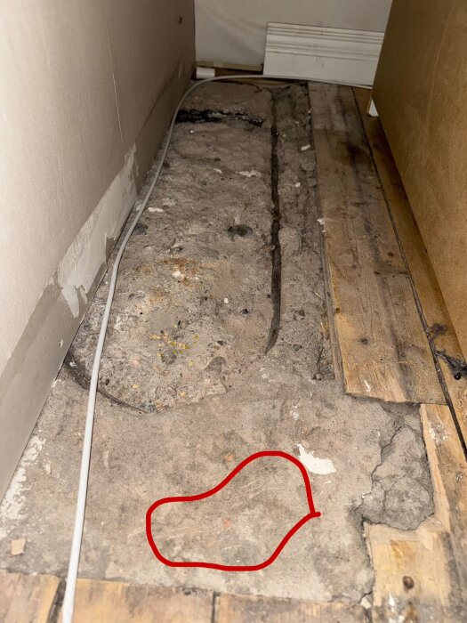 Bilden visar en del av ett rum där golvet är uppbrutet med exponerad betong och trägolv. En kabel ligger längs väggen och en röd markering syns på betongytan.