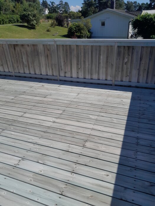 En väl underhållen träterrass och ett staket syns i en solig trädgårdsmiljö, omgivna av gräs och en vit byggnad i bakgrunden.