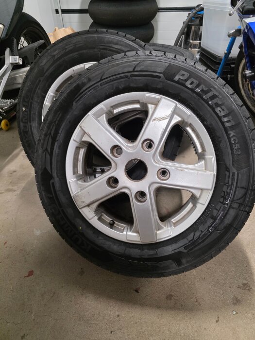 Två nya bildäck med fälgar på ett garagegolv, redo för montering inför säsongens däckbyte. Bakgrund av verktyg och gamla däck.