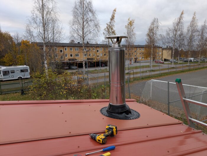 Skorsten monterad på ett rött plåttak med verktyg i förgrunden, omgiven av träd och byggnader i bakgrunden.