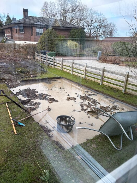 Översvämmad utgrävd grop med lerigt vatten, en skottkärra och rensgaller. Bakgrund med gräs och staket framför ett hus.