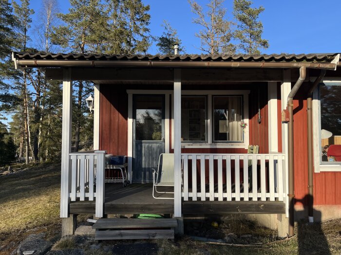 Röd sommarstuga med liten altan, vita staket och en enkel uppsättning utemöbler i soligt väder.