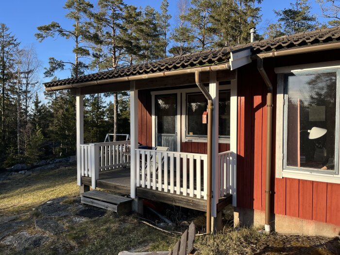 Altan vid rött sommarhus med trätak och vita räcken, omgiven av träd, planeras att byggas om till innerum, soligt väder.