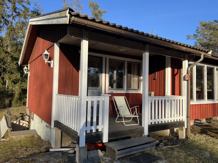 Röd sommarstuga med altan, vit räcke och solstol. Altanen har stödpelare och stege. Huset är omgivet av träd och gräsmatta.