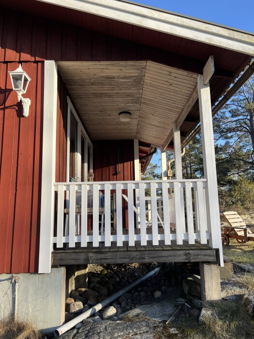 Röd sommarstuga med liten altan upphöjd på plintar, vit staketbalkong, gräsmatta bredvid och utemöbler skymtar i bakgrunden.
