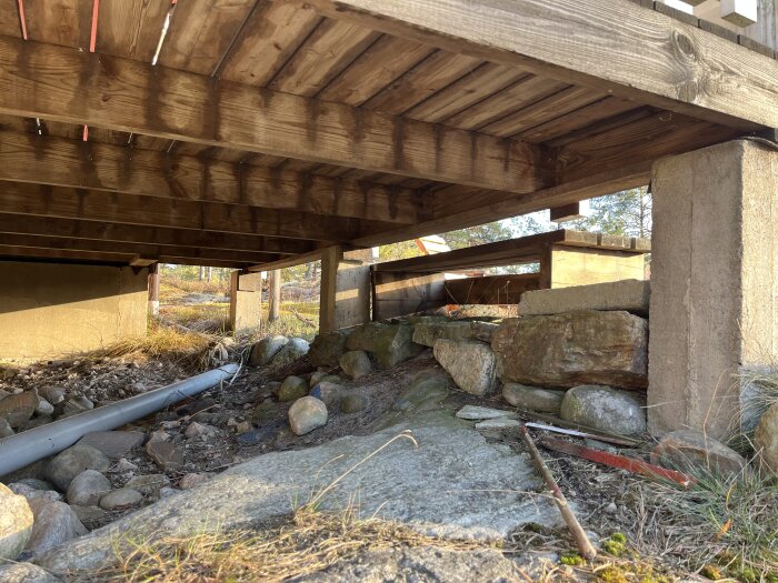 Altanen sedd underifrån med träbalkar, betongplintar och stenar. Visar befintlig grund för planerad ombyggnad till innerum.