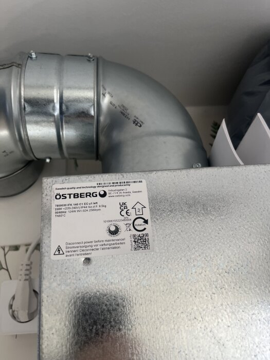 Ventilationsrör och en rektangulär metallbox med etikett från Östberg. Boxen har märkningar och en QR-kod.