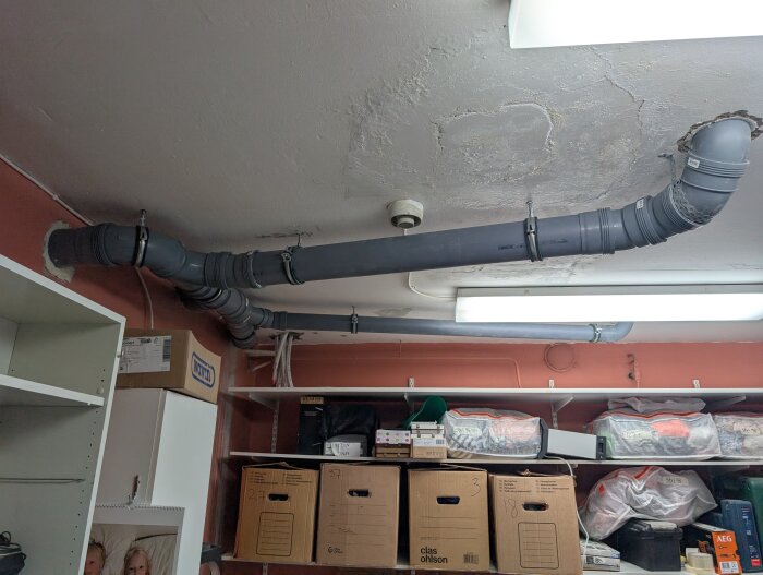 Grå avloppsrör i plast installerade i taket av ett rum med hyllor fyllda med kartonger och plastlådor.