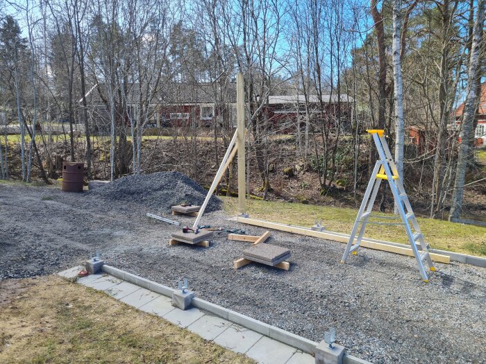 Första stolpen rest i byggprojekt, omgiven av grus och en stege, med verktyg och ritningar på marken. Bakgrund av träd och röd trähus.