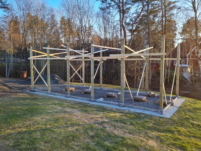 Trästomme under uppbyggnad på en grusad yta med skog och hus i bakgrunden. Leverans av byggmaterial nyligen anlänt.