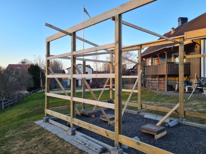 Trästomme under uppbyggnad i en trädgård, med flera resade stolpar och horisontella reglar. Hus och gräsmatta syns i bakgrunden.