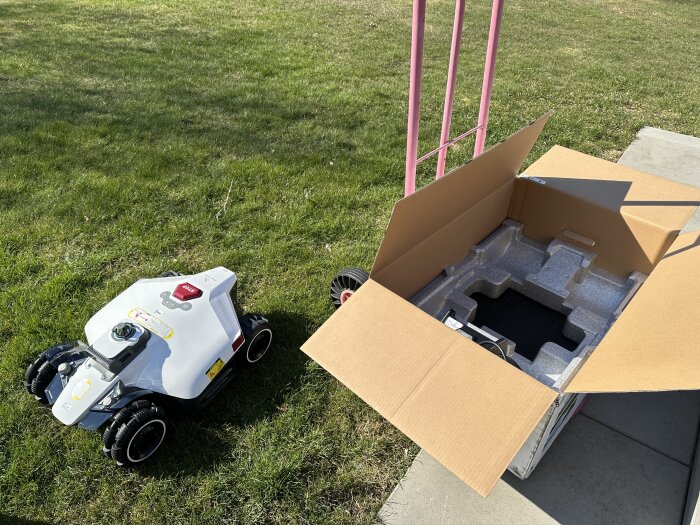 Robotgräsklippare bredvid en öppen kartong på en gräsmatta i solen.
