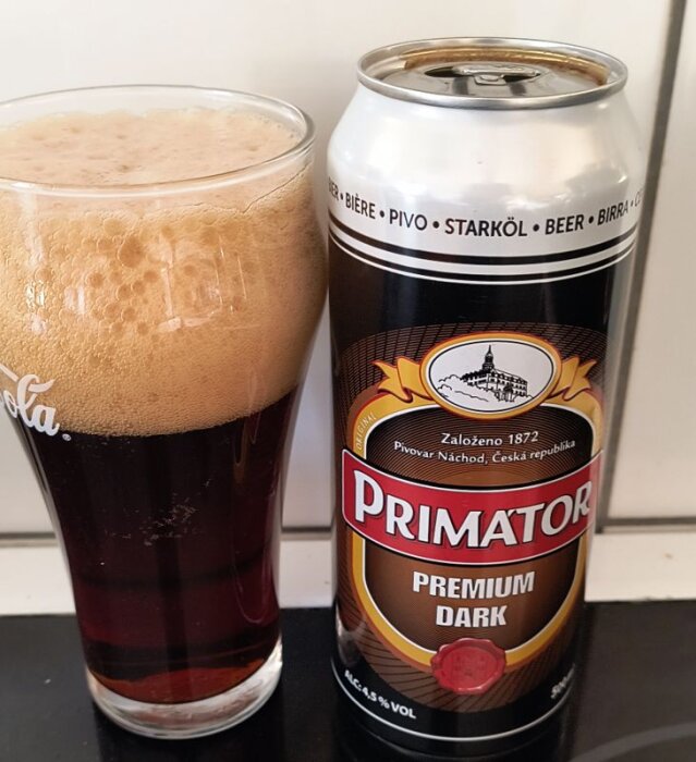 En burk Primator Premium Dark öl bredvid ett glas med skummande mörk öl.